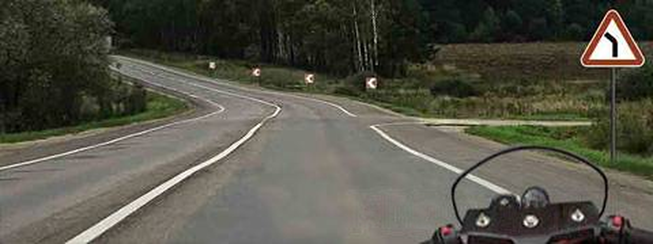 Motorcycle view of a road curving left through trees; a red triangle shows a black bend symbol.